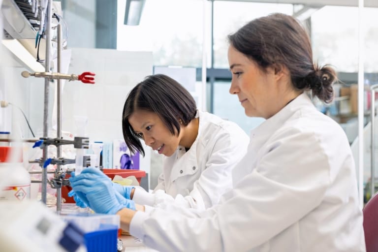 Kelly Nguyen at work in her biomedical research laboratory, next to a lab colleague. They are wearing white laboratory coats and blue gloves.